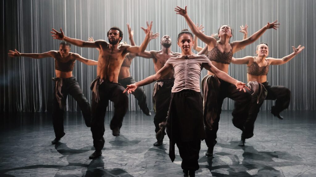 The full company advance towards the audience with arms outstretched, in front of a wall of silver strands.